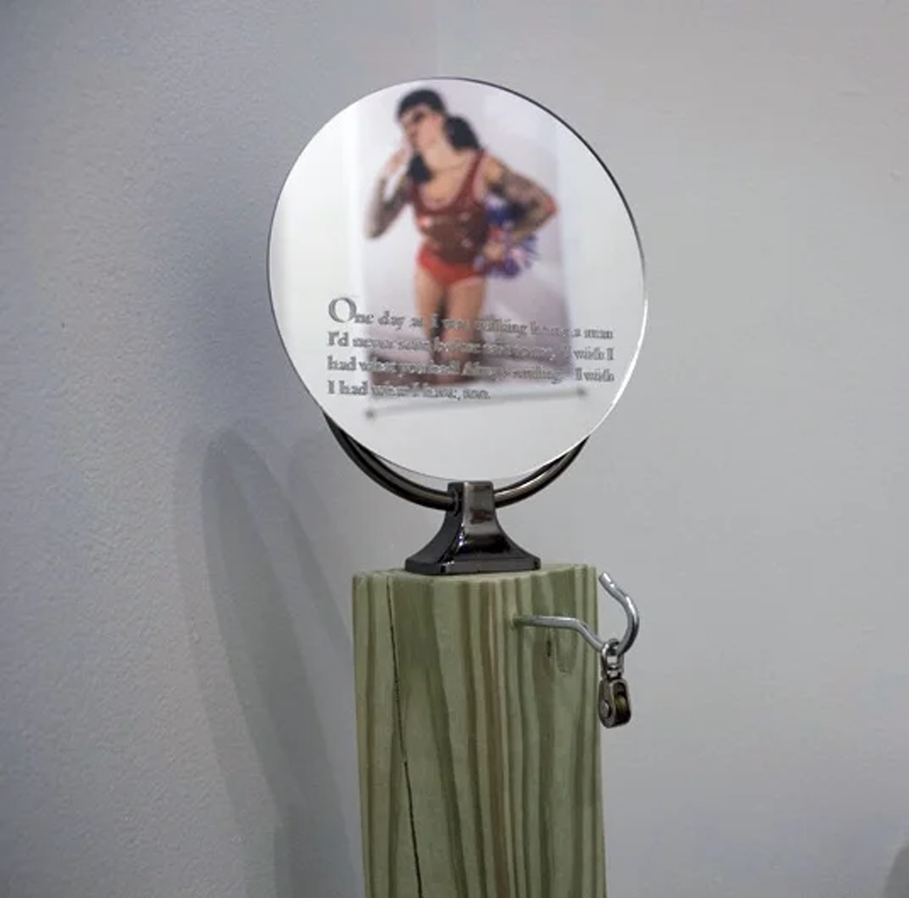 Installation photograph of EchoLocation exhibition. Photograph by Heather Raquel Phillips of the artist smoking a cigarette in a cheerleader costome is reflected off sculpture by Sarah Legow of a mirror with text etched into it mounted to a wooden post. 