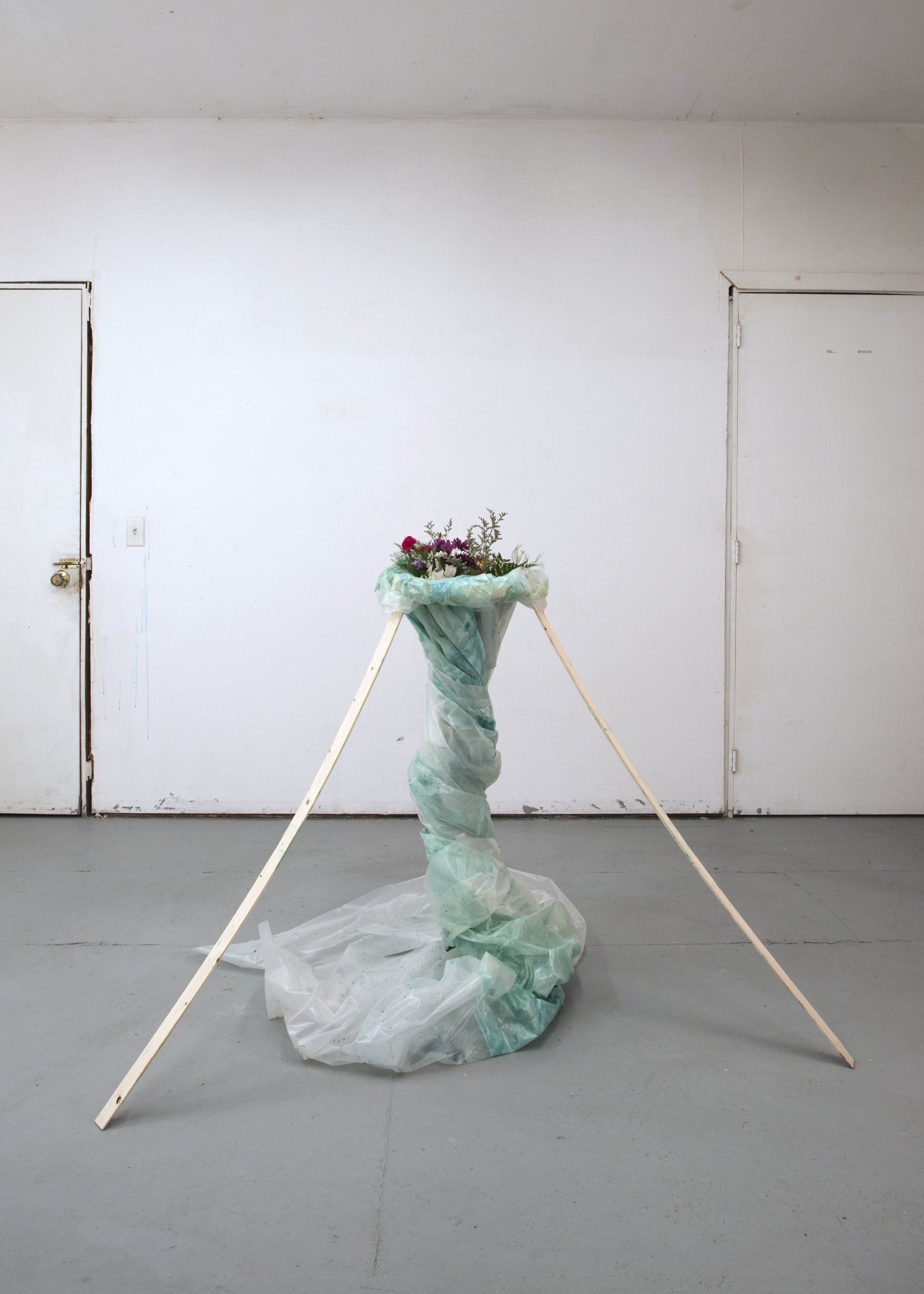 Sculpture of toilet bowl cleaner stained plastic wrap gathered into a vortex shape propped up by three thin wooden strips and with a plastic boquet emerging from the vortex mouth at the top.