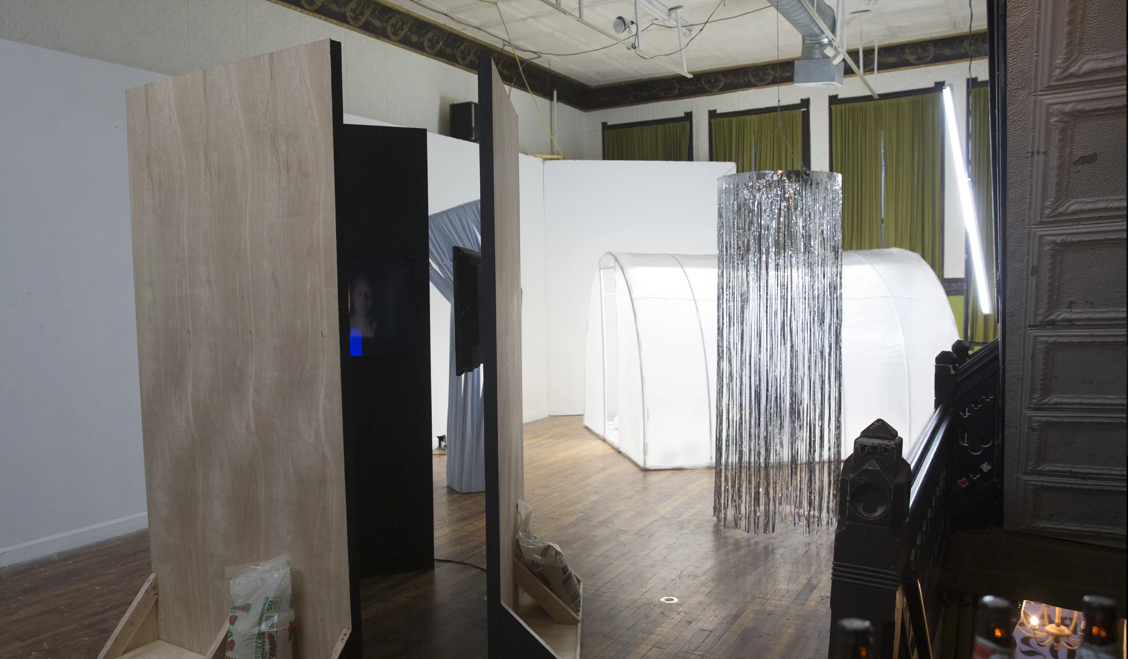 Installation photograph of Please Come In. A freestanding sculpture by Daria McMeans of three walls painted black with TV screens mounted on each and facing each other; sit in front of a hanging kinetic sculpture by Keenan Jackson of tinsel arranged in a circle slowly spinning, and another hanging sculpture by the same artist of flourescent lights; in front of a freestanding scultpure by Stephanie Elden of a hoop house illuminated from within and containing cast plaster cabbages.