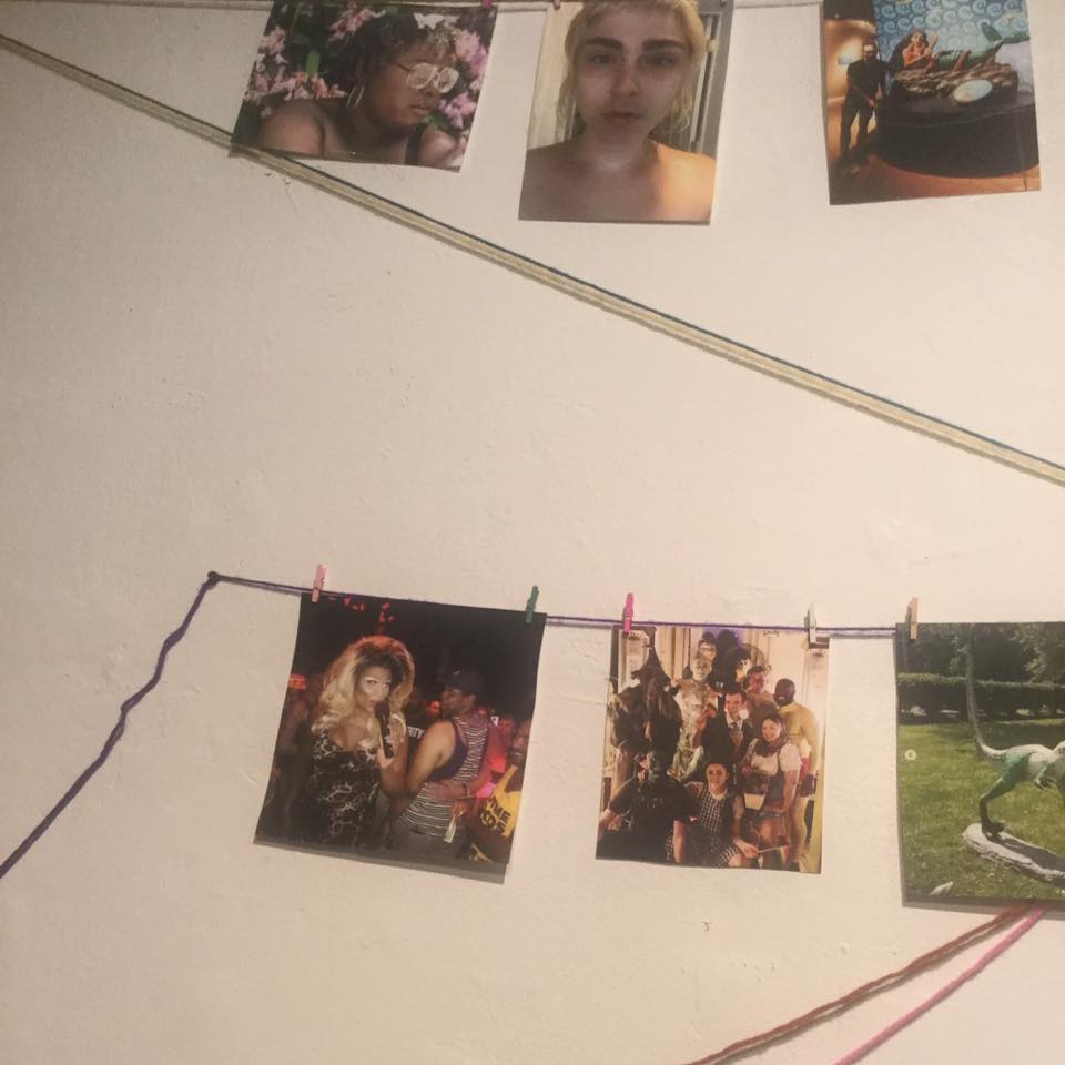 Installation photograph of exhibition submissions clothespinned to colored yarn stretched across gallery wall.