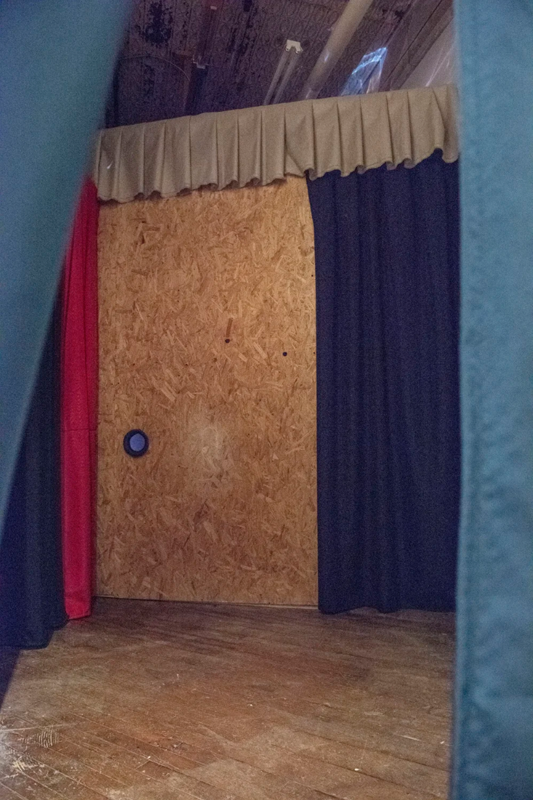 Installation photograph through curtain into space with a plywood wall with a hole cut out in the lower middle, the wall flanked by curtains on either side.