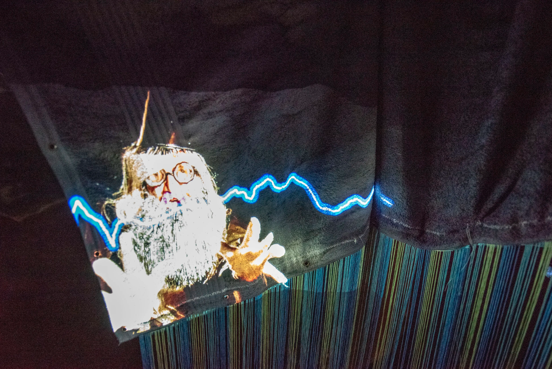 Installation photograph showing video of wizard character projected on a hanging towel with blue fringe attached below.