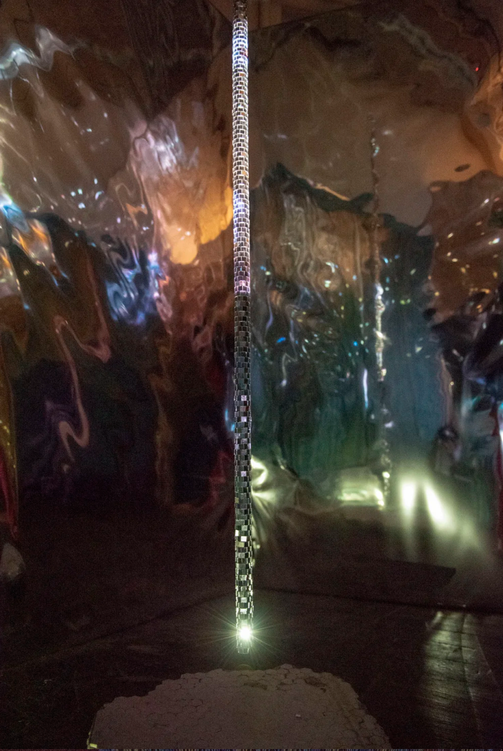 Installation photograph of disco-mirrored rod hovering above chunk of broken tile floor with lit mylar in the background.
