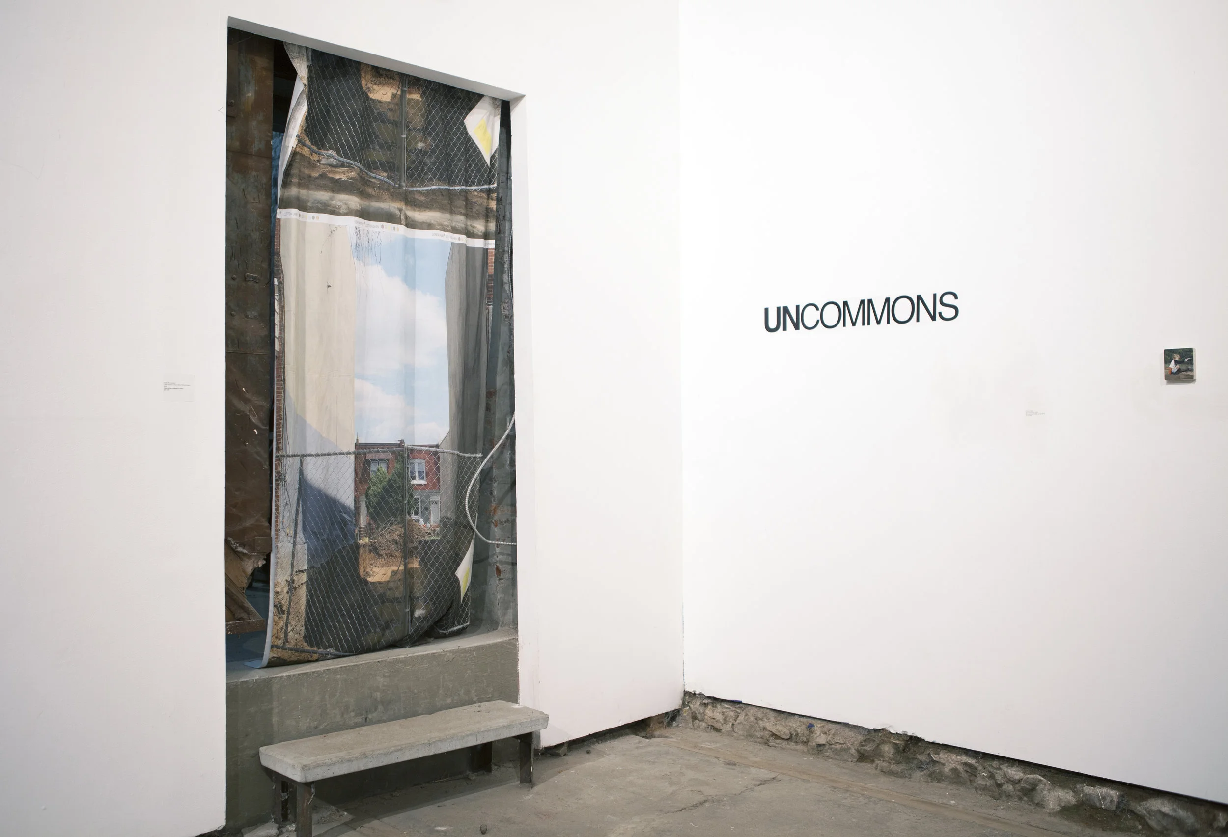 Installation photograph of Uncommons exhibition. A Kaitlin Pomerantz artwork in form of curtain with a photo of a gentrifying cityscape printed on it, hanging in a doorway, adjacent to Uncommons wall text.
