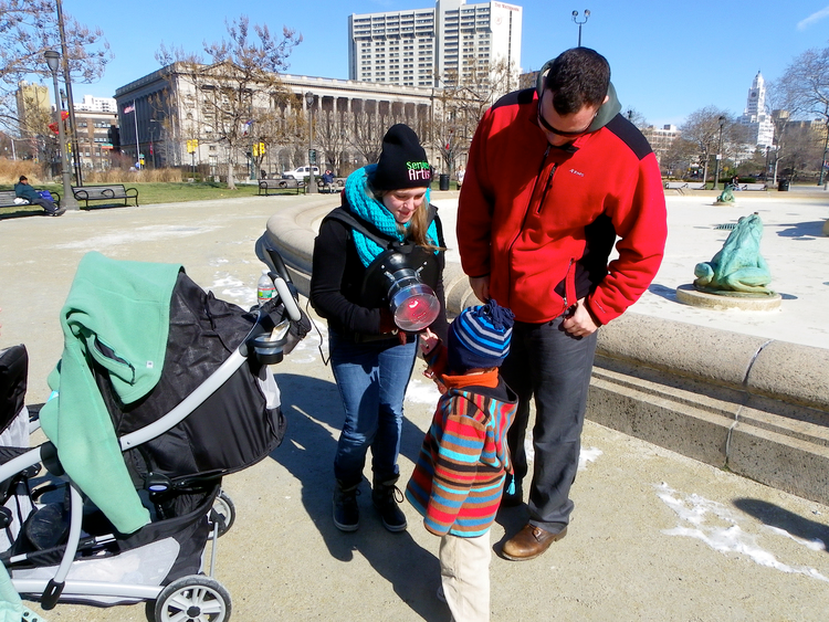 Service artist providing warmth using heat lamp harness to young viewer beside fountain.